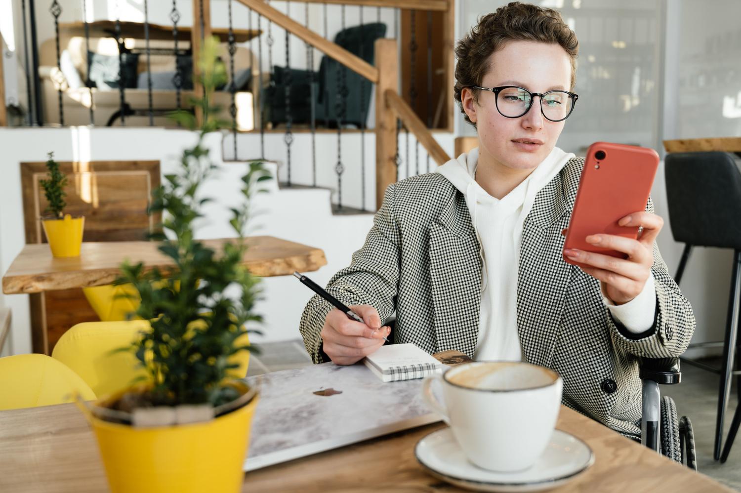 Mujer profesional en silla de ruedas trabajando con su teléfono móvil en una cafetería moderna.