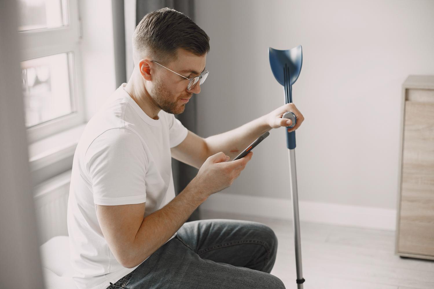 Hombre utilizando muletas y un smartphone en el hogar, ilustrando la accesibilidad tecnológica.