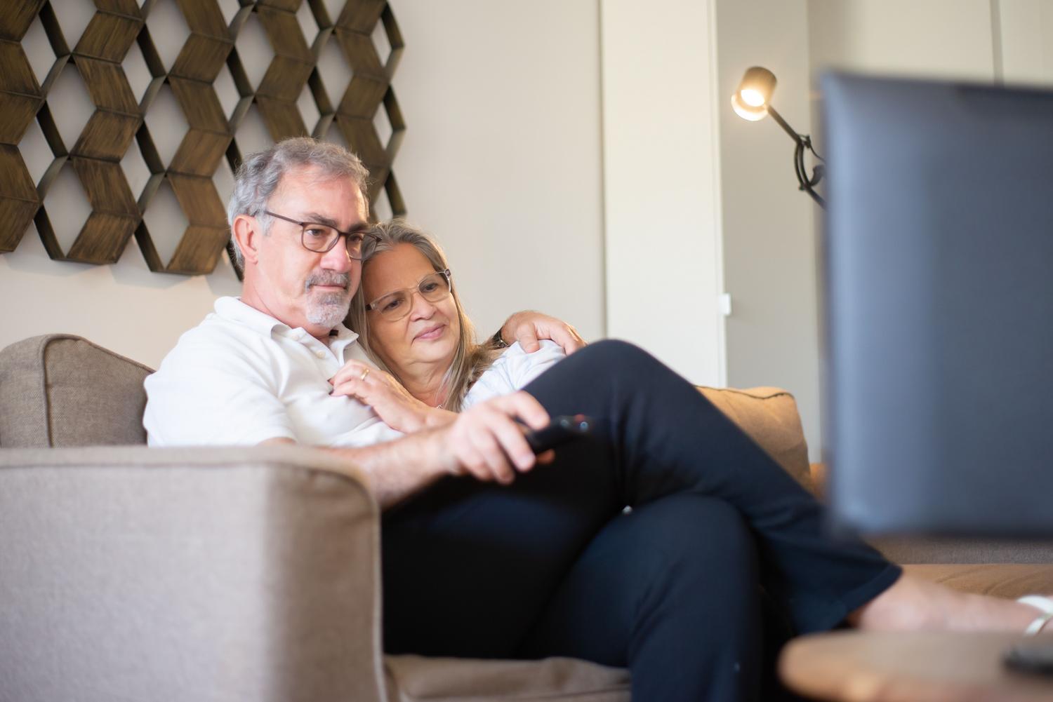 Pareja de adultos mayores disfrutando de la televisión en su hogar, ilustrando la inclusión tecnológica.