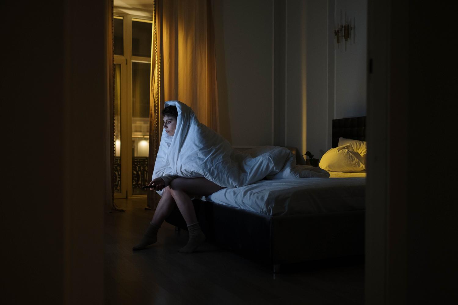 Mujer envuelta en una manta viendo la televisión en un dormitorio oscuro, ilustrando el impacto de la luz azul en el sueño.