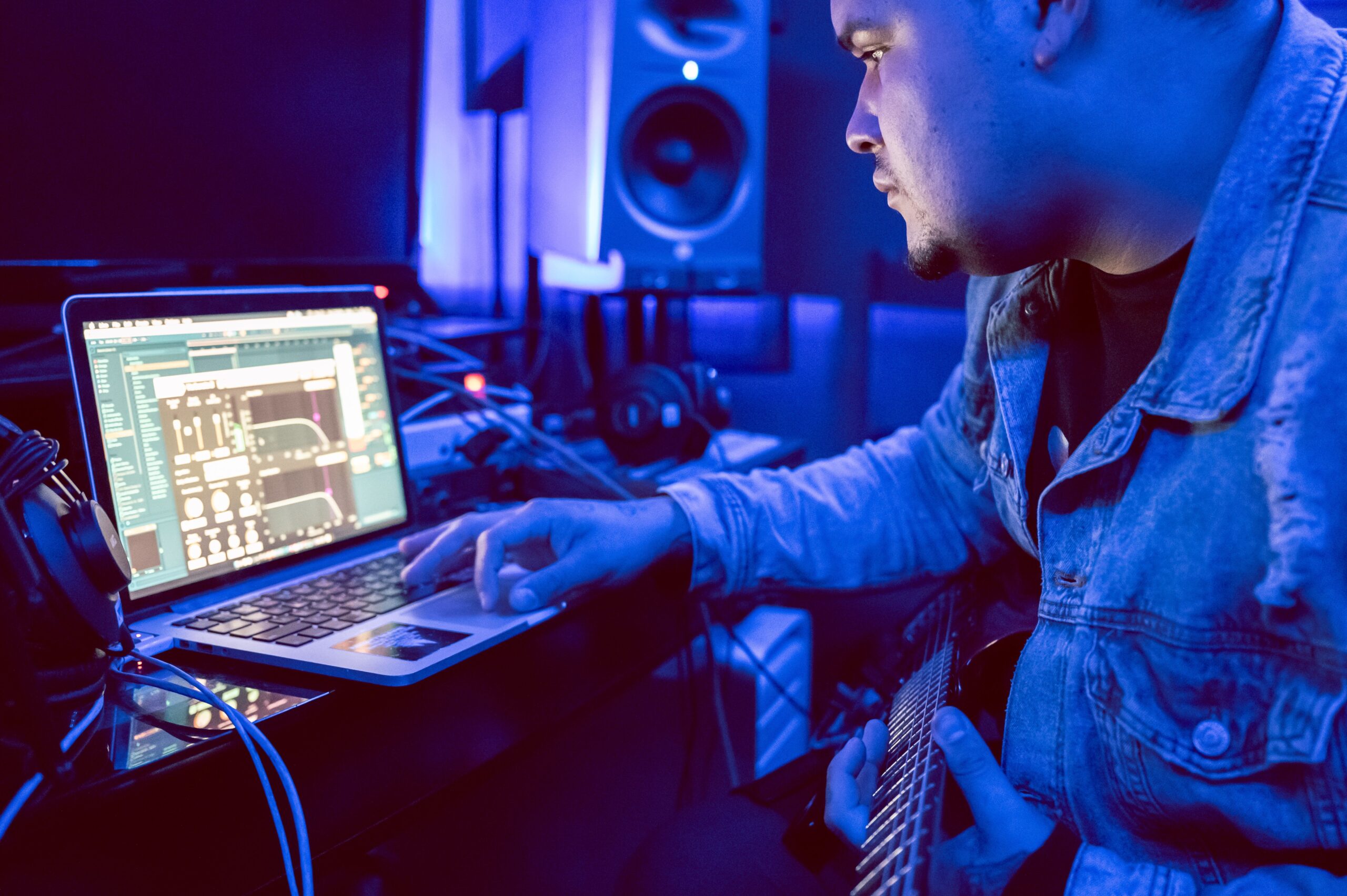 Músico en un estudio de grabación utilizando una laptop y guitarra eléctrica bajo una iluminación azul, representando la necesidad de baja latencia al grabar.