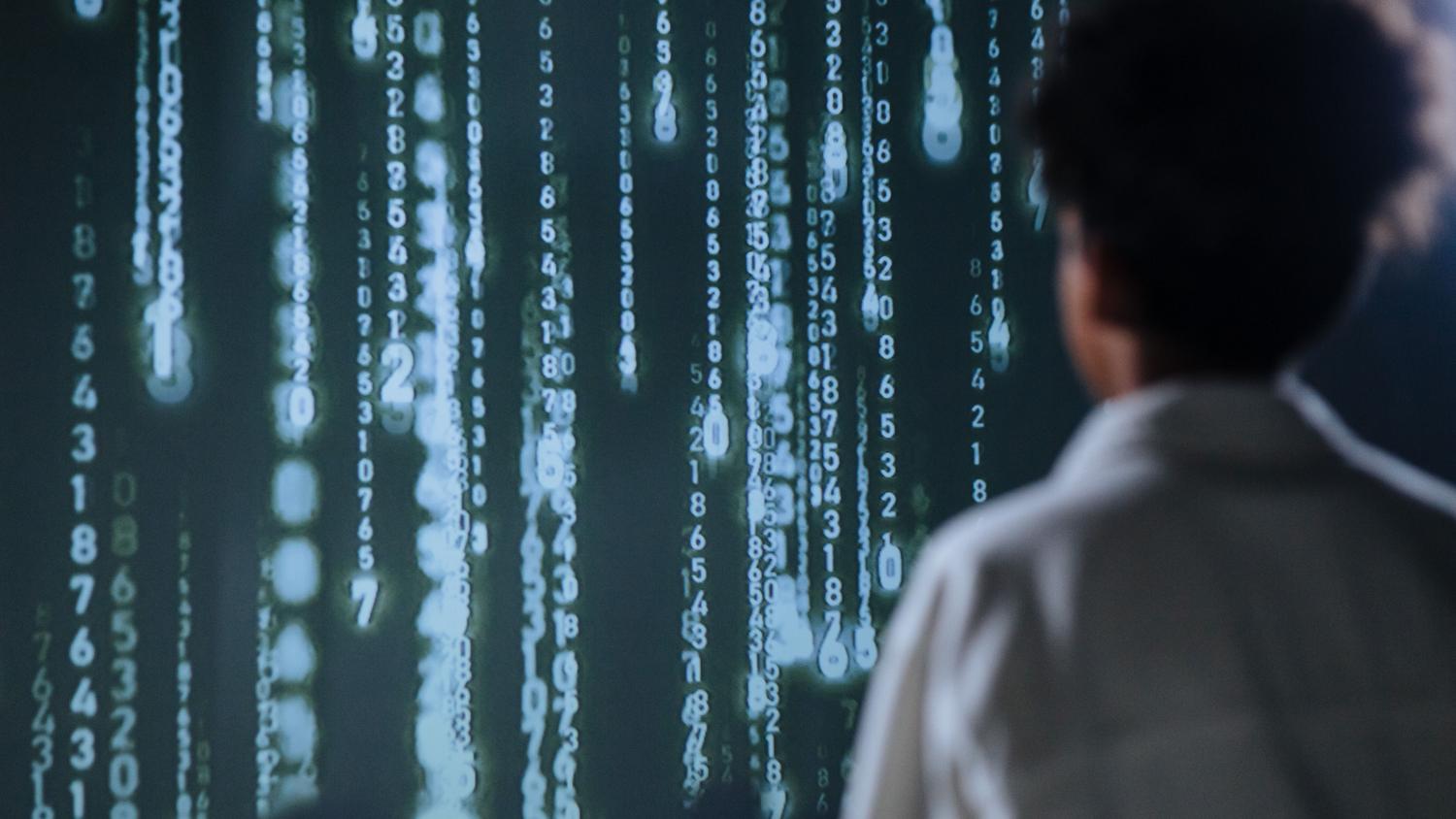 Persona analizando flujos de datos y números en una pantalla gigante, evocando la capacidad de procesamiento y automatización de tareas complejas.