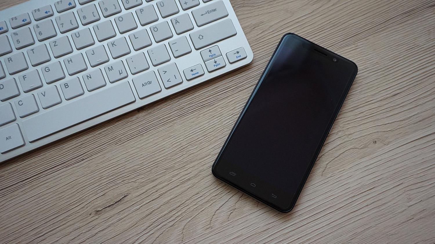 Primer plano de un smartphone junto a un teclado en un escritorio de madera, representando la sincronización del ecosistema de dispositivos.