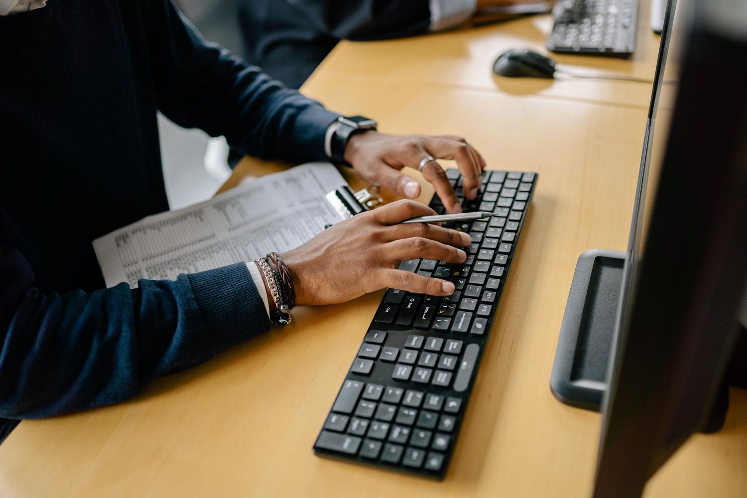 Primer plano de manos tecleando en un teclado de ordenador sobre un escritorio de madera, ilustrando la productividad y el trabajo técnico.