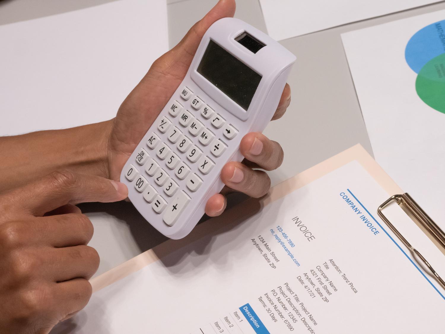Manos utilizando una calculadora junto a una factura de empresa, ilustrando el proceso de cálculo de impuestos y totales en la facturación.