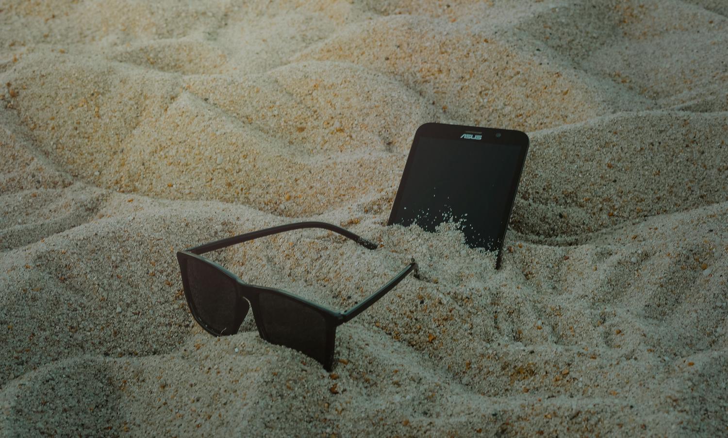 Smartphone y gafas de sol sobre la arena de la playa, representando la exposición peligrosa del dispositivo al sol en verano.