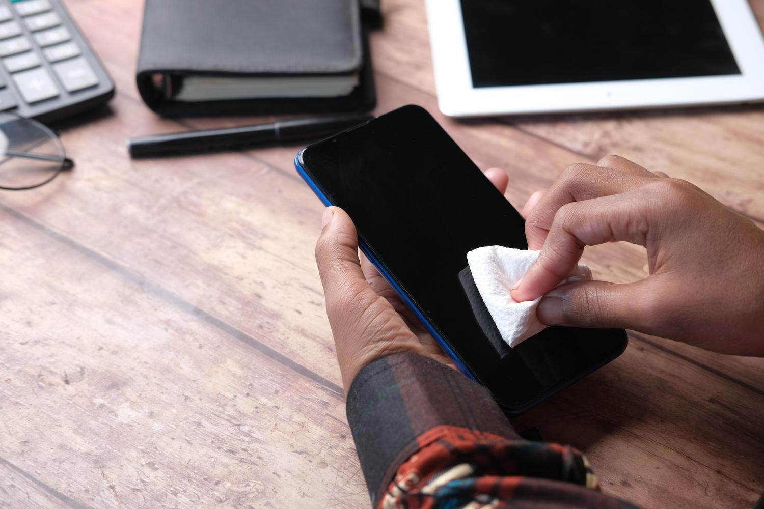 Manos limpiando la pantalla de un teléfono inteligente en un escritorio organizado, transmitiendo una sensación de mantenimiento profesional.