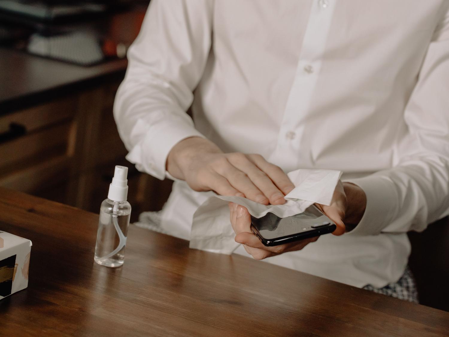 Persona desinfectando la pantalla de un smartphone utilizando un paño y un pulverizador con solución limpiadora sobre una mesa de madera.