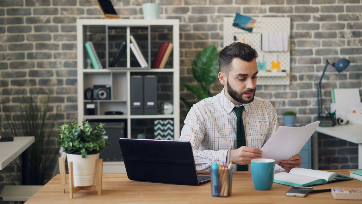 Hombre de negocios concentrado leyendo informes extensos en su escritorio con un portátil al lado.