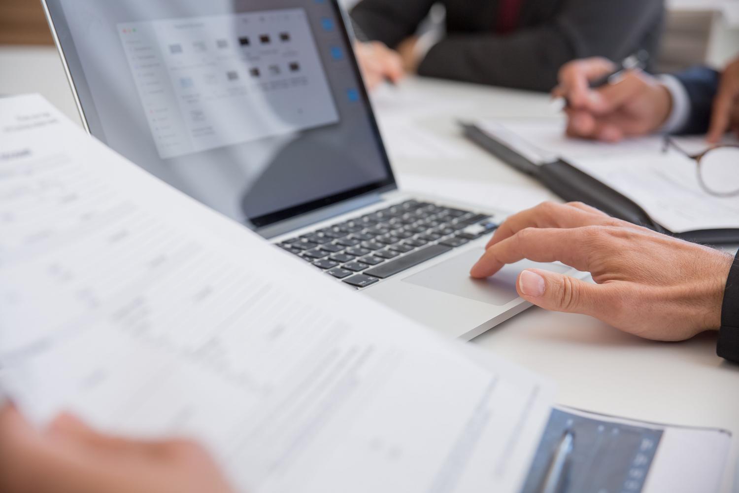 Primer plano de manos trabajando con documentos y un ordenador portátil, ilustrando el flujo de trabajo y la productividad.