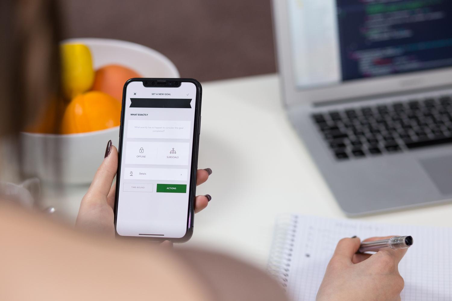 Persona utilizando un iPhone y tomando notas en un ordenador portátil en un espacio de trabajo moderno, representando la productividad con IA.