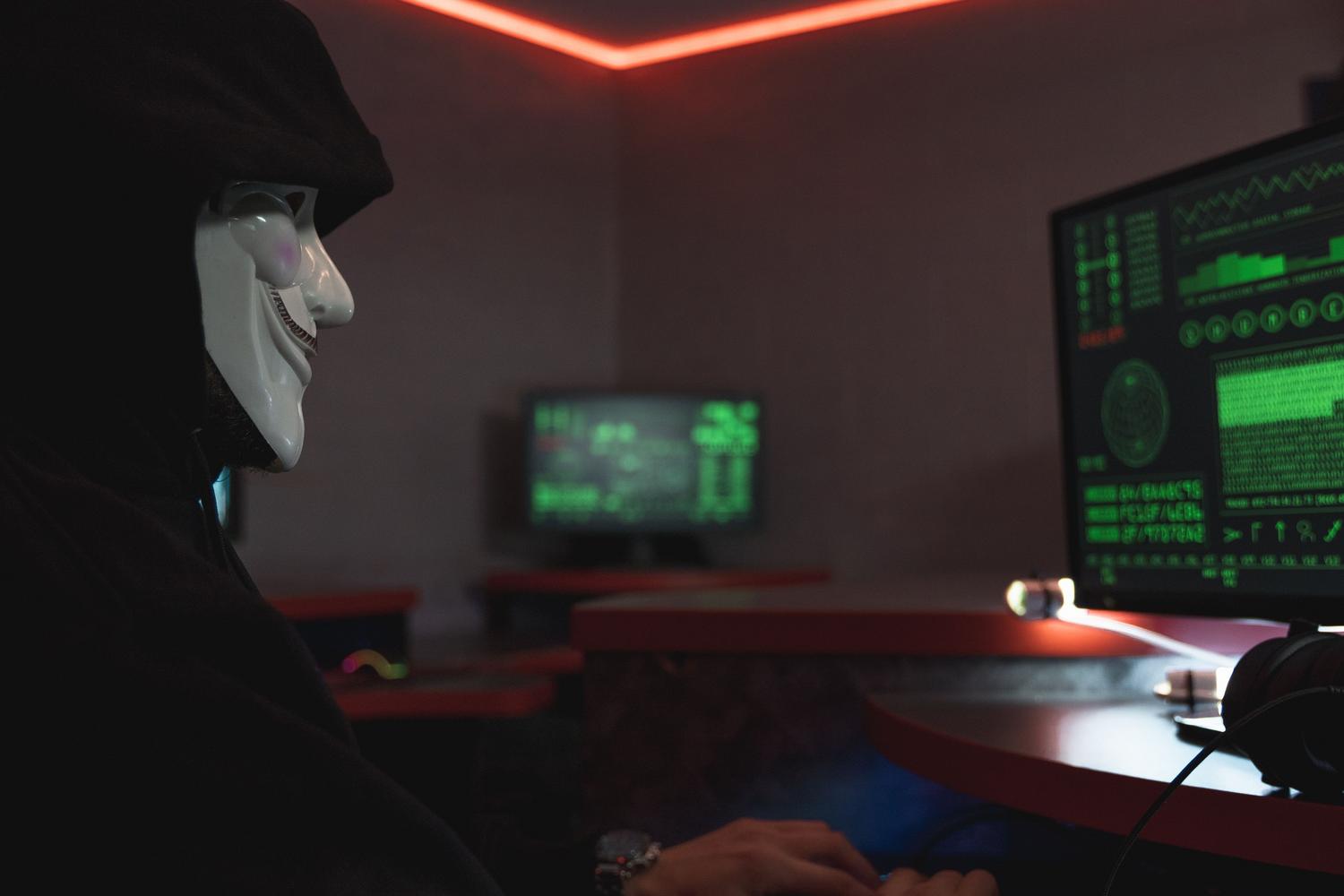 Hacker con máscara de Guy Fawkes operando ordenadores en un entorno clandestino.