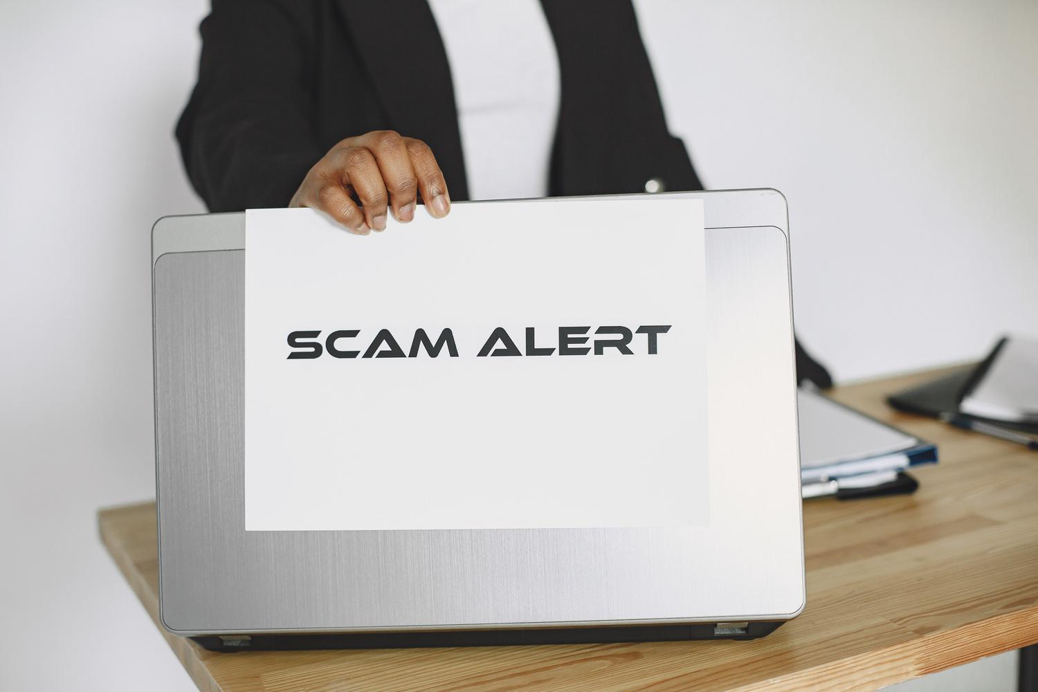 Persona sosteniendo un cartel de 'SCAM ALERT' sobre un portátil, ilustrando las alertas de fraude y phishing.