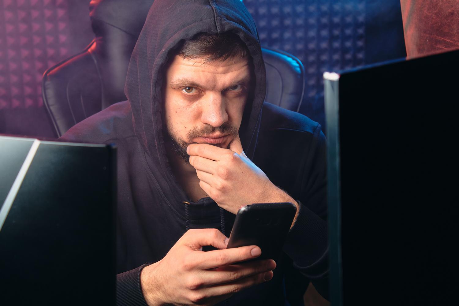 Hombre con capucha utilizando un smartphone en un entorno tecnológico oscuro, representando a un ciberdelincuente.