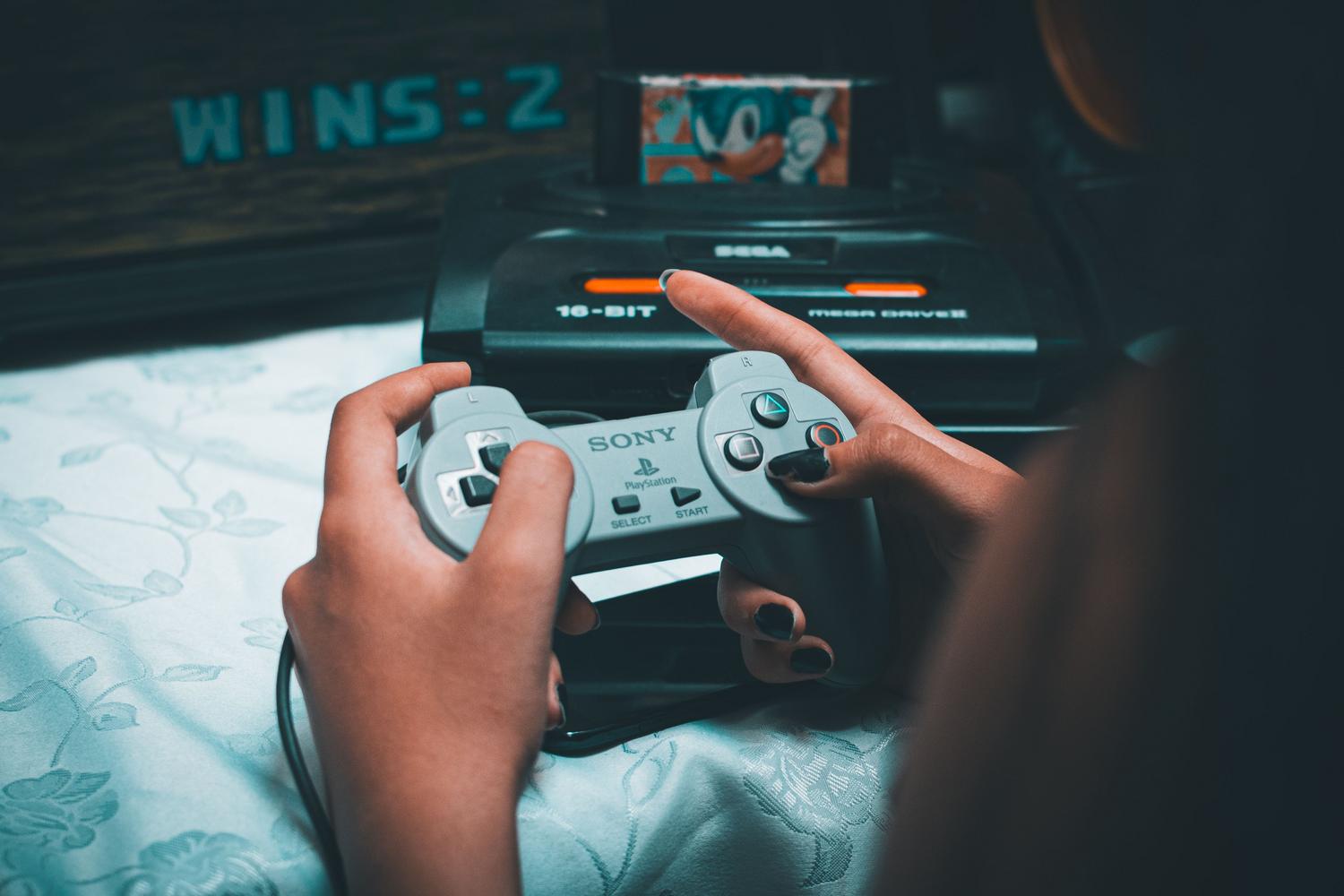 Manos sosteniendo un mando vintage de Sony con una Sega Mega Drive al fondo.