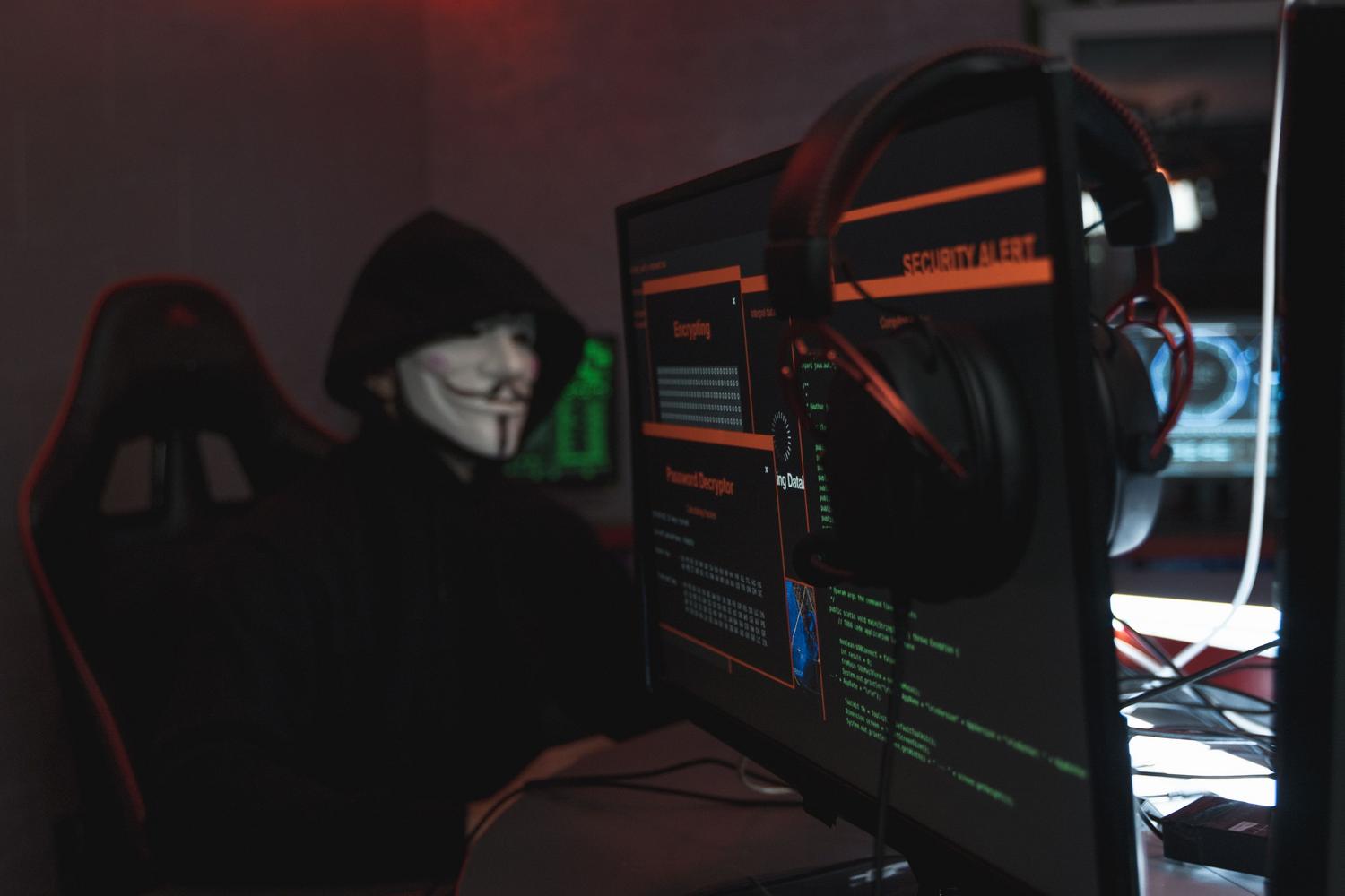 Ciberdelincuente con máscara de Anonymous operando un sistema informático en un entorno oscuro, representando la amenaza del malware y los ataques dirigidos